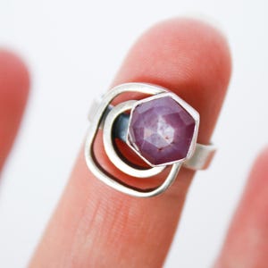 May include: A silver ring featuring a faceted, hexagonal, purplish-red gemstone. The ring's design includes a geometric, openwork setting with a square shape and two concentric circles around the gemstone.