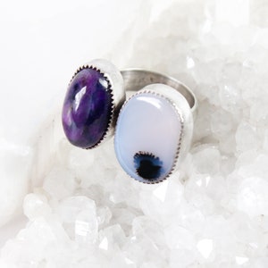 May include: Two silver rings with oval-shaped gemstones. The ring on the left has a purple gemstone, and the ring on the right has a white gemstone with a black and blue swirl.