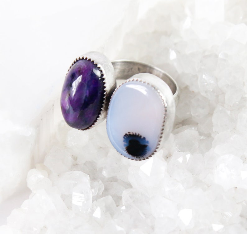 May include: Two silver rings with oval-shaped gemstones. The ring on the left has a purple gemstone, and the ring on the right has a white gemstone with a black and blue swirl.