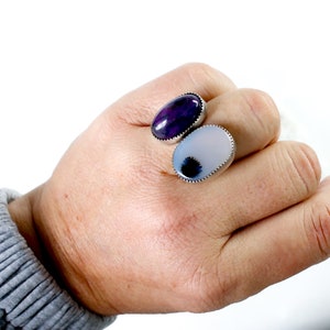May include: Two oval-shaped rings with silver settings. The ring on the left has a purple stone with white veins. The ring on the right has a white stone with a black center.