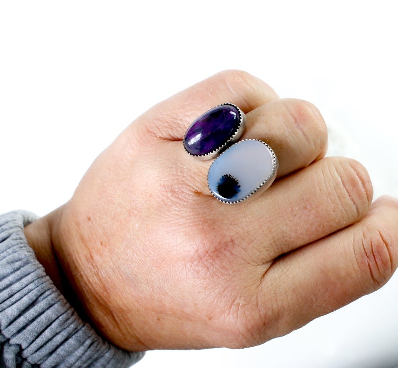 May include: Two oval-shaped rings with silver settings. The ring on the left has a purple stone with white veins. The ring on the right has a white stone with a black center.