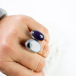 May include: Two oval-shaped rings with silver settings. The ring on the left has a white agate stone with black markings. The ring on the right has a purple agate stone.