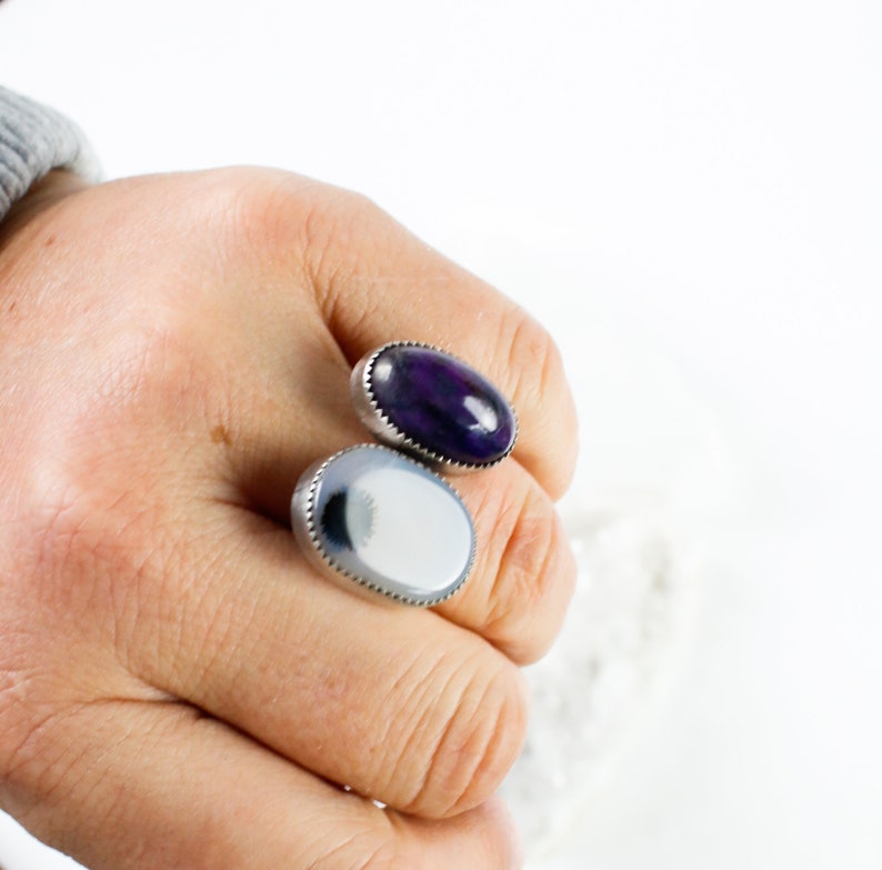 May include: Two oval-shaped rings with silver settings. The ring on the left has a white agate stone with black markings. The ring on the right has a purple agate stone.