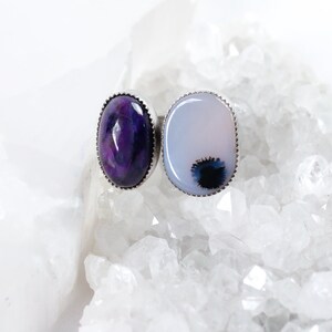 May include: Two oval-shaped rings with silver settings. The ring on the left has a purple and white stone. The ring on the right has a white stone with a black and gray pattern.