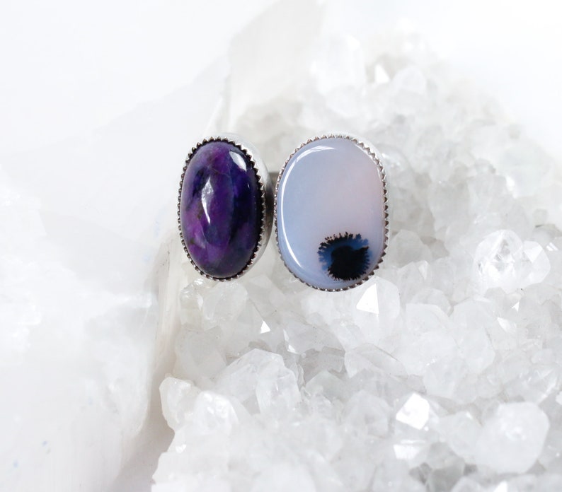 May include: Two oval-shaped rings with silver settings. The ring on the left has a purple and white stone. The ring on the right has a white stone with a black and gray pattern.