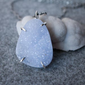 May include: A silver chain necklace with a light blue agate pendant. The pendant is a freeform shape and has a sparkly, crystalline texture.