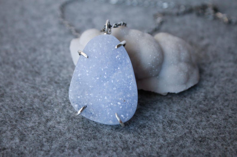 May include: A silver chain necklace with a light blue agate pendant. The pendant is a freeform shape and has a sparkly, crystalline texture.