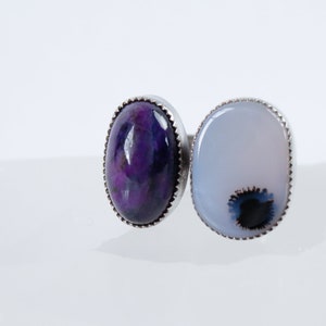 May include: A silver ring with two oval-shaped gemstones. The left stone is a deep purple with swirls of color. The right stone is a light blue with a dark blue and black swirl in the center.