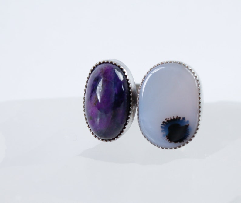 May include: A silver ring with two oval-shaped gemstones. The left stone is a deep purple with swirls of color. The right stone is a light blue with a dark blue and black swirl in the center.