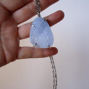 May include: A silver chain necklace with a light blue, teardrop-shaped pendant. The pendant is made of a natural stone with a sparkly, textured surface.
