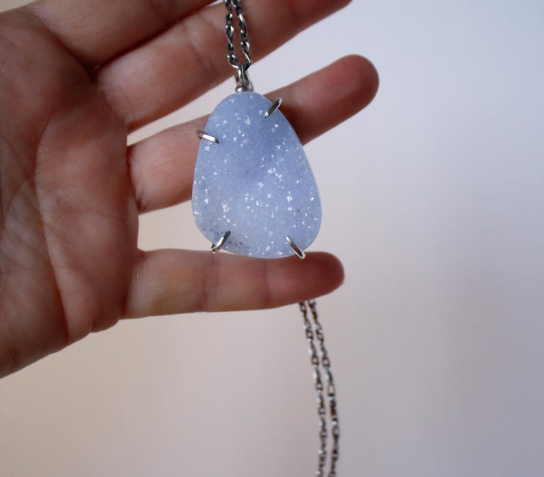 May include: A silver chain necklace with a light blue, teardrop-shaped pendant. The pendant is made of a natural stone with a sparkly, textured surface.