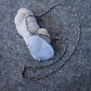 May include: A silver chain necklace with a light blue, irregularly shaped stone pendant. The stone is set in a silver wire frame.