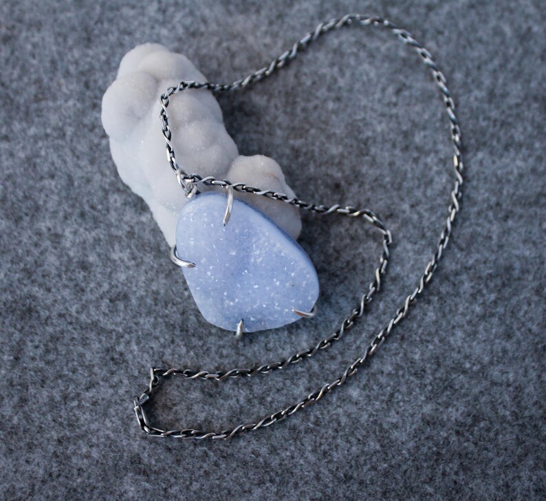 May include: A silver chain necklace with a light blue, irregularly shaped stone pendant. The stone is set in a silver wire frame.