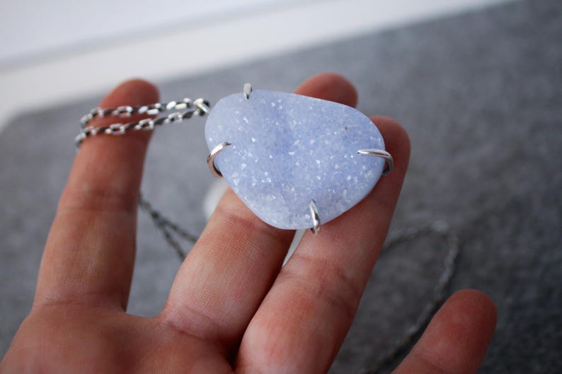 May include: A silver chain necklace with a light blue agate pendant. The pendant is a freeform shape and has a sparkly, crystalline texture.