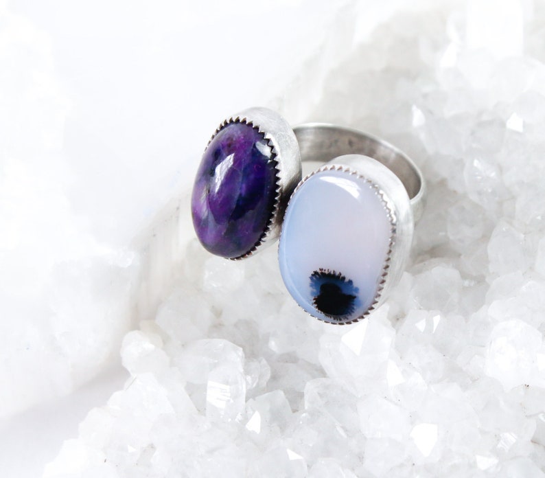 May include: A silver ring with two oval-shaped gemstones. The left stone is a deep purple with white and gray swirls. The right stone is a light blue with a dark gray center.