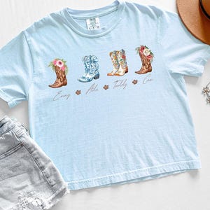 May include: Light blue t-shirt featuring four watercolor cowboy boots with floral accents. Each boot has a different design and is labeled with a name. The shirt is paired with denim shorts and a brown hat.