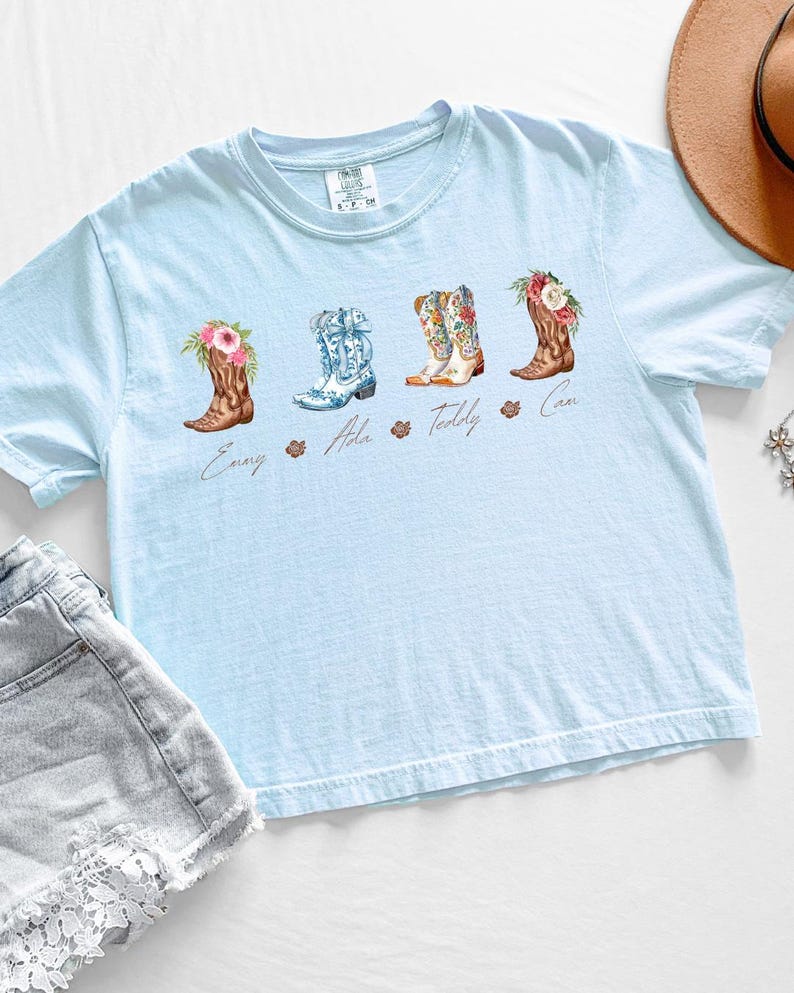 May include: Light blue t-shirt featuring four watercolor cowboy boots with floral accents. Each boot has a different design and is labeled with a name. The shirt is paired with denim shorts and a brown hat.