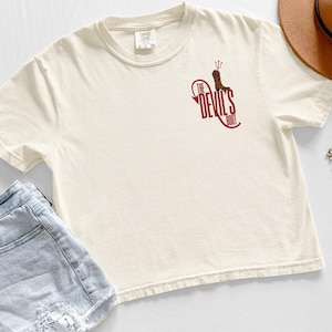 May include: Cream-colored t-shirt with a maroon graphic that reads "The Devil's Boot" and features a boot with a trident. The shirt is short-sleeved and has a relaxed fit. The shirt is paired with denim shorts and a brown hat.