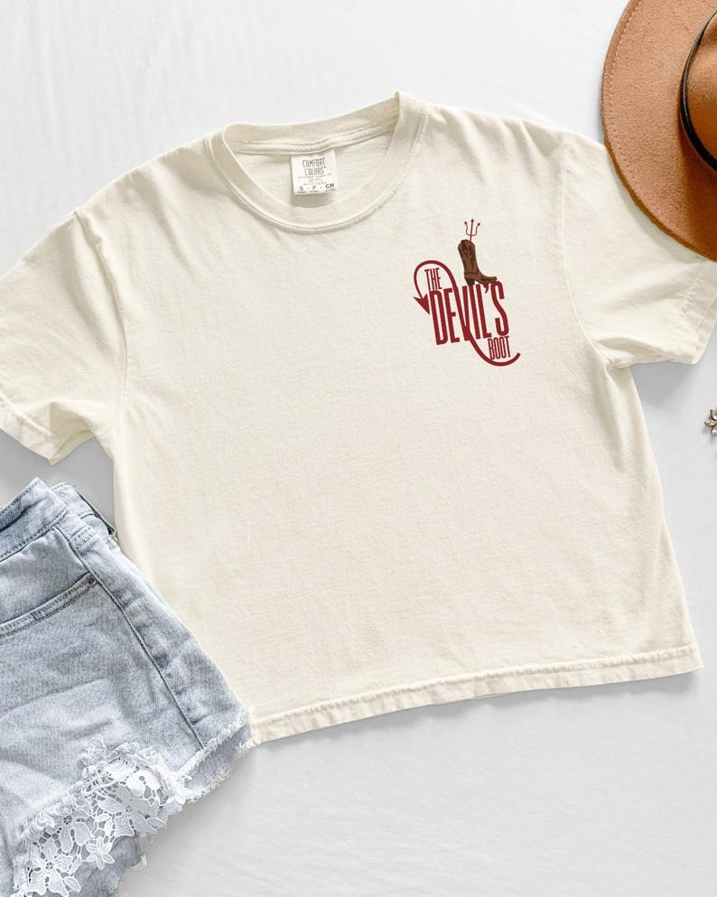 May include: Cream-colored t-shirt with a maroon graphic that reads "The Devil's Boot" and features a boot with a trident. The shirt is short-sleeved and has a relaxed fit. The shirt is paired with denim shorts and a brown hat.