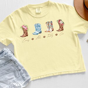 May include: A light yellow t-shirt featuring four watercolor cowboy boots with floral accents. Each boot has a different design and color scheme, with names below each boot. The shirt is paired with denim shorts and a brown hat.