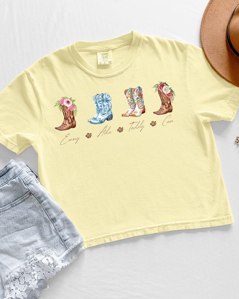 May include: A light yellow t-shirt featuring four watercolor cowboy boots with floral accents. Each boot has a different design and color scheme, with names below each boot. The shirt is paired with denim shorts and a brown hat.