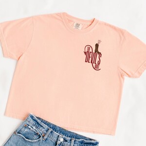 May include: A peach-colored t-shirt with the text "The Devil's Boot" in maroon. The graphic includes a boot and a pitchfork. Light blue denim jeans are visible below the shirt.