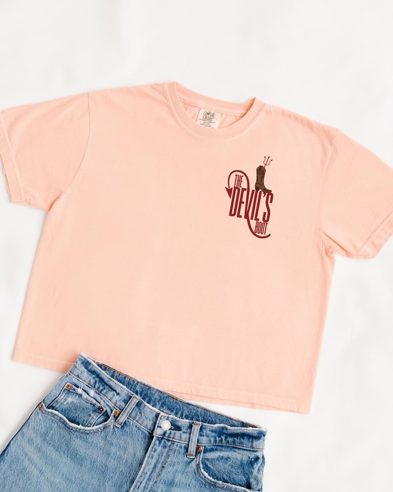 May include: A peach-colored t-shirt with the text "The Devil's Boot" in maroon. The graphic includes a boot and a pitchfork. Light blue denim jeans are visible below the shirt.