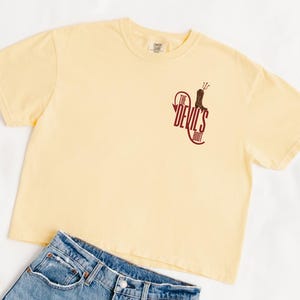 May include: A light yellow t-shirt with the text "The Devil's Boot" in maroon. The shirt is paired with blue denim jeans. The t-shirt has a crew neck and short sleeves. The graphic on the shirt features a cowboy boot.