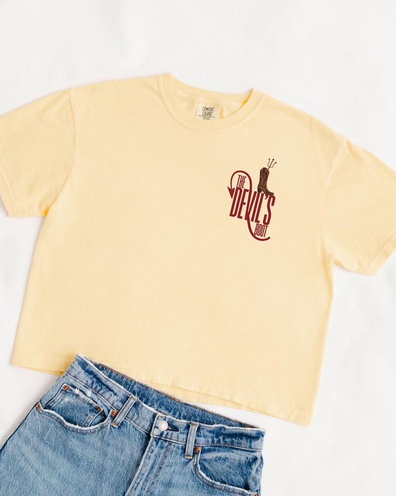 May include: A light yellow t-shirt with the text "The Devil's Boot" in maroon. The shirt is paired with blue denim jeans. The t-shirt has a crew neck and short sleeves. The graphic on the shirt features a cowboy boot.
