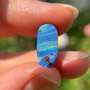 Pode incluir: Uma gema de opala oval, exibindo uma gama de azuis, verdes e tons dourados. A pedra polida é segurada entre os dedos, destacando seus padrões naturais. As cores da opala mudam com a luz, criando um efeito visual dinâmico. Mede aproximadamente 2 cm de comprimento.
