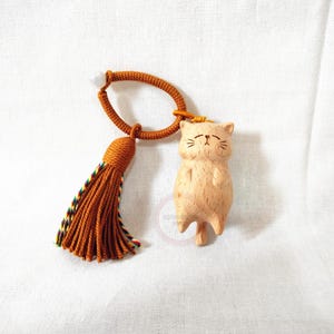 May include: A wooden cat figurine with a tassel and cord. The cat is light brown with etched facial features. The tassel is orange with a multicolored trim. The cord is orange and forms a loop. The background is white.