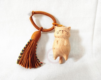 May include: A wooden cat figurine with a tassel and cord. The cat is light brown with etched facial features. The tassel is orange with a multicoloured trim. The cord is orange and forms a loop. The background is white.
