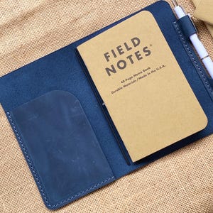 May include: A blue leather notebook cover with a Field Notes memo book inside. A white pen is secured in a leather loop. The memo book is tan with black text that reads "FIELD NOTES". The cover has a pocket on the left side.