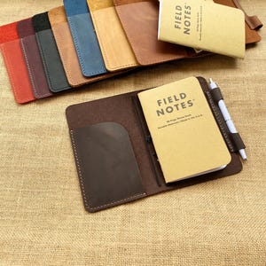May include: A collection of leather Field Notes journal covers in various colors, including red, black, blue, and tan. Each cover holds a small notebook with the "Field Notes" logo. A pen is secured in a loop on one of the covers. The items are arranged on a burlap surface.