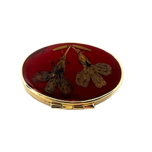 May include: Oval compact with a gold-tone metal frame and a deep red enamel top. The top features two dried flowers. The compact is closed with a small clasp.