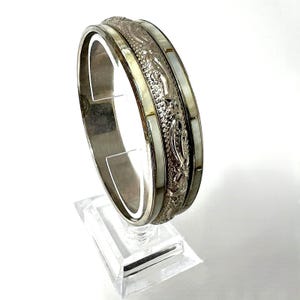 Può includere: Bracciale rigido argentato con dettagli in rilievo ornati e sezioni intarsiate di madreperla bianca. Il bracciale è esposto su un supporto in acrilico trasparente, che mette in risalto il suo design intricato. Un gioiello classico.