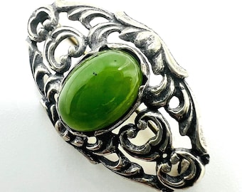 May include: A silver-toned ring featuring an oval, bright green gemstone. The ring has an ornate, openwork design with swirling patterns around the stone. The gemstone is the focal point, set against the detailed metalwork.