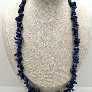 May include: A necklace made of dark blue gemstone chips, each with an irregular shape. The chips are strung together to form a continuous loop. The necklace is displayed on a mannequin with a neutral-coloured fabric.