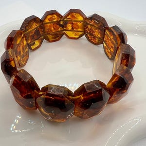 Könnte beinhalten: Ein facettiertes Bernsteinarmband in warmem Honigbraun. Das Armband besteht aus großen, geometrisch geformten Perlen, die ein auffälliges und einzigartiges Design bilden. Die Perlen sind zu einem flexiblen, tragbaren Accessoire aufgereiht.