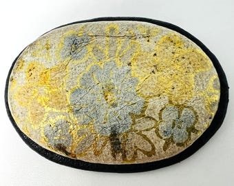 Könnte beinhalten: Ovales Dekorationselement mit schwarzem Rand. Die Mitte hat eine strukturierte Oberfläche mit einem Blumenmuster in Gold-, Grau- und Beigetönen. Das Design hat eine Vintage- oder Antik-Ästhetik.