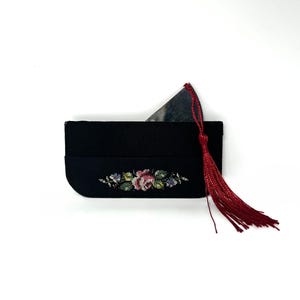 Könnte beinhalten: Schwarze Clutch mit floraler Stickerei in Rosa, Grün und Blau. Ein silbernes Objekt ist teilweise im Inneren sichtbar. Eine lange rote Quaste hängt an der Seite. Die Clutch befindet sich auf weißem Hintergrund.