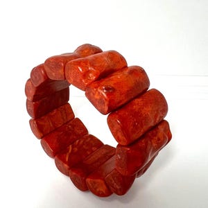Könnte beinhalten: Ein rot-oranges Armband aus rechteckigen, polierten Perlen. Die Perlen sind zu einer flexiblen, kreisförmigen Form aufgereiht. Das Armband hat ein natürliches, strukturiertes Aussehen. Durchmesser ca. 7 cm.