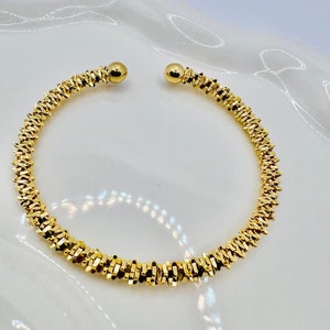 Può includere: Un braccialetto a polsino aperto color oro. Il braccialetto ha un design strutturato e sfaccettato ed è aperto alle estremità, ciascuna sormontata da una piccola perla rotonda dorata. Il braccialetto è esposto su una superficie bianca.