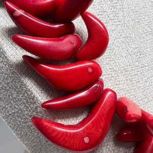 May include: Close-up of a red coral necklace. The necklace features large, curved, polished coral beads. The beads are a vibrant red color and have a smooth, glossy finish. The necklace is displayed on a light gray textured surface.