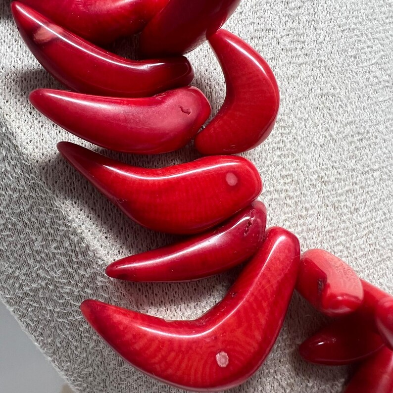 May include: Close-up of a red coral necklace. The necklace features large, curved, polished coral beads. The beads are a vibrant red color and have a smooth, glossy finish. The necklace is displayed on a light gray textured surface.