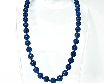 May include: A beaded necklace featuring deep blue, spherical beads. The necklace is strung on a dark cord and secured with a silver-colored clasp. The beads have a speckled appearance, and the necklace is displayed on a clear stand.