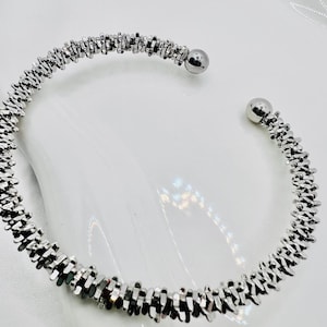 Può includere: Un bracciale a polsino color argento con un design strutturato e segmentato. Il bracciale ha un design aperto e flessibile con una piccola sfera rotonda ad ogni estremità. Il bracciale è mostrato su una superficie bianca.