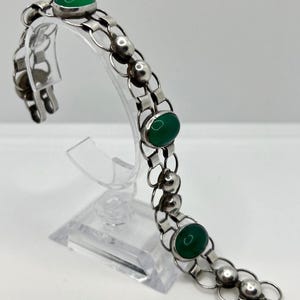 Puede incluir: Una pulsera de plata con tres piedras preciosas verdes ovaladas y una serie de eslabones y esferas plateadas. La pulsera se exhibe sobre un soporte transparente sobre un fondo blanco. La pulsera es una joya.