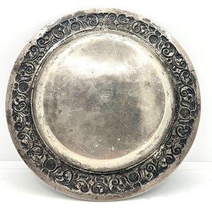 Könnte beinhalten: Ein runder, silberfarbener Teller mit einem verzierten, erhabenen floralen und Rankenwerk-Rand. Die Mitte des Tellers ist glatt und weist Gebrauchsspuren auf. Der dekorative Rand verleiht diesem Servierteller eine Vintage-Ästhetik.