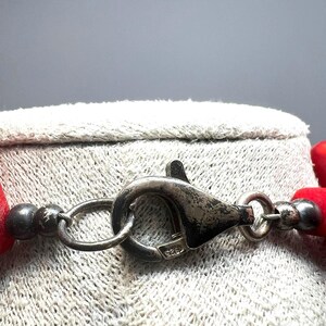 May include: Close-up of a silver necklace clasp with a lobster claw closure. The clasp is connected to a strand of red beads. The clasp is stamped with "925". The necklace is resting on a textured, light gray surface.